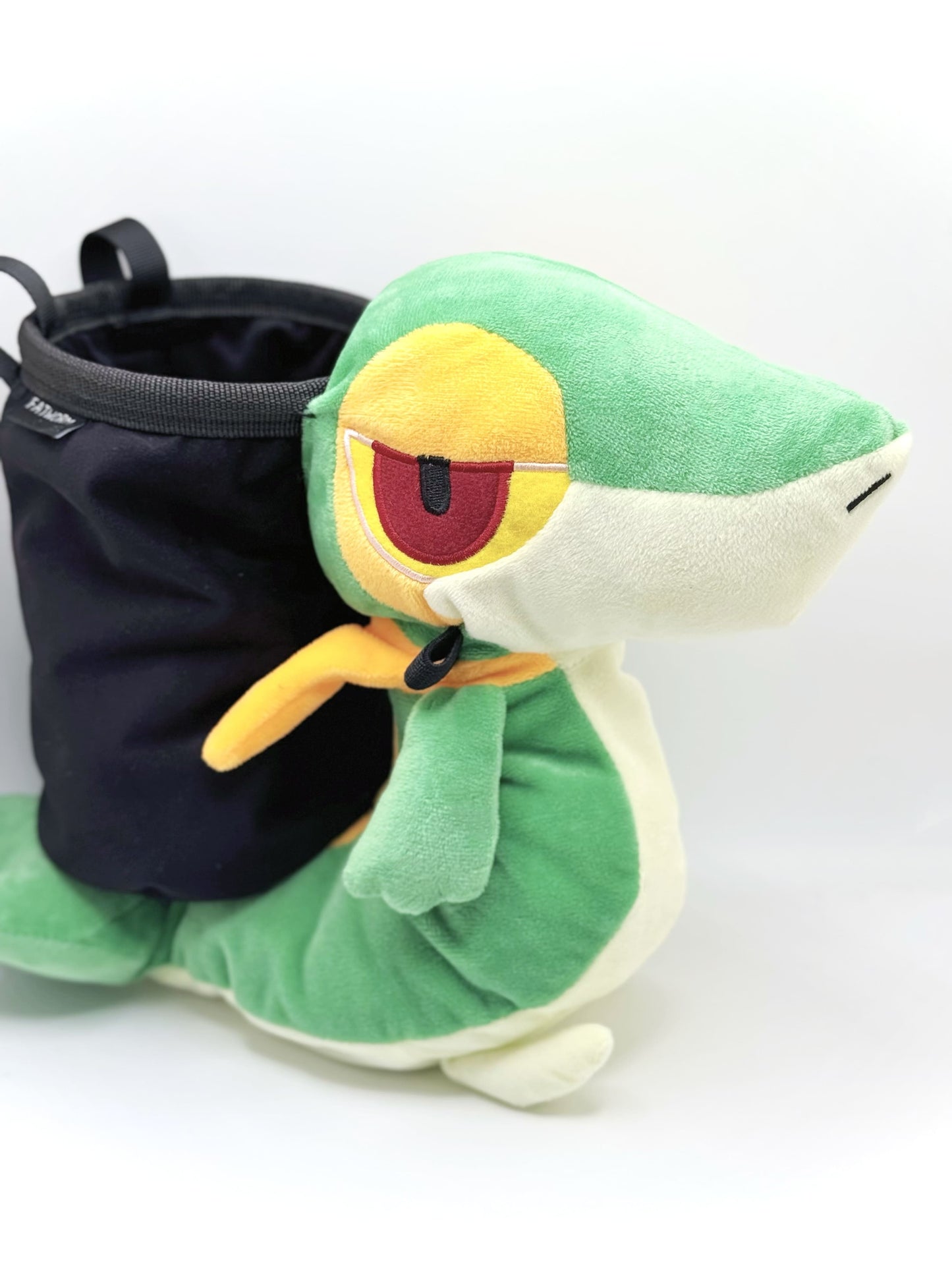 A cute, handmade plush climbing chalk bag designed to look like the Snivy, featuring a sleek green and cream body with yellow leaf-like accents and large, confident eyes. Designed with high functionality by a climber, it includes a wide black rim opening at the top for easy chalk access. A great climbing gift for trainers who want to catch them all.