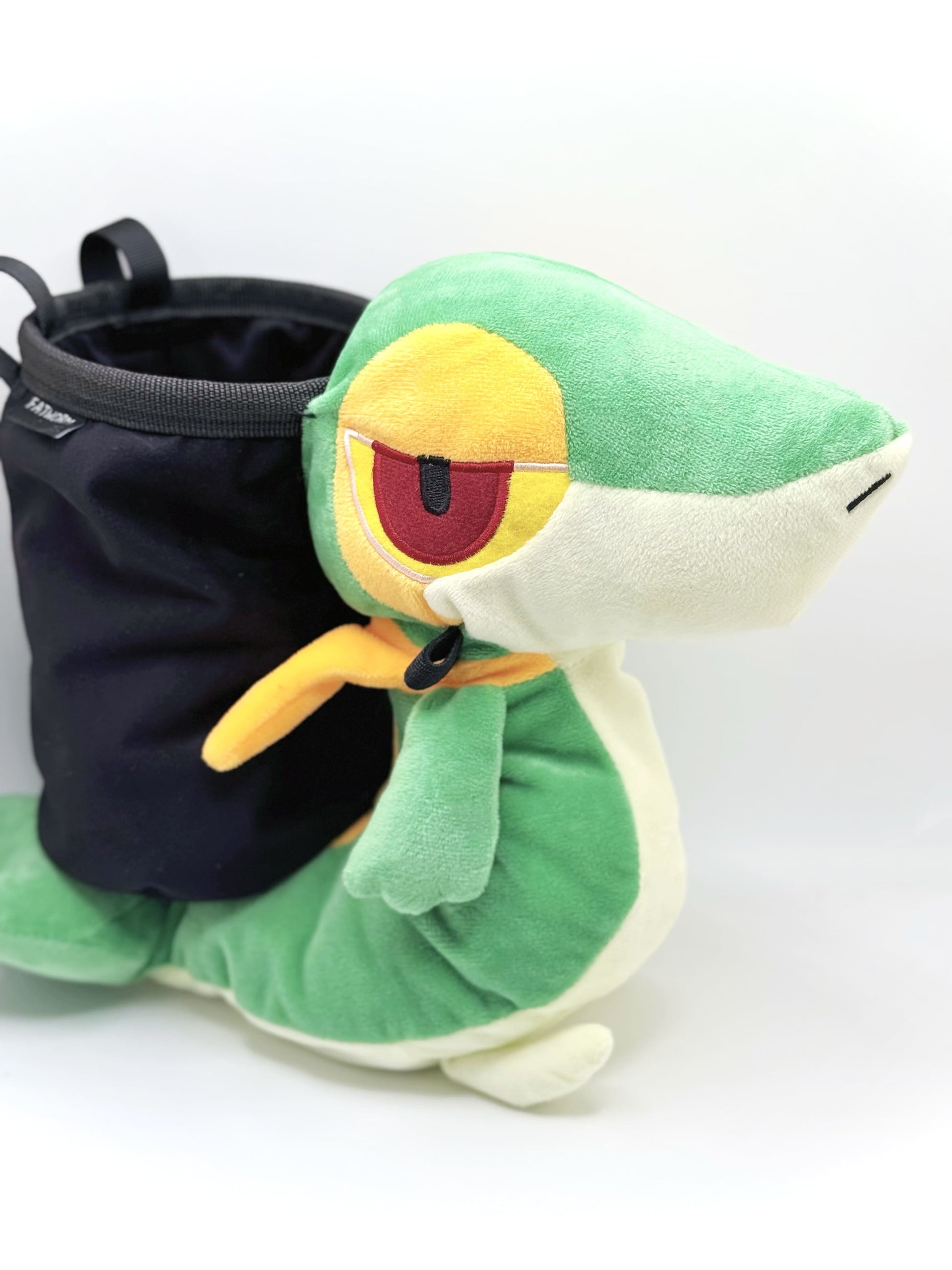 A cute, handmade plush climbing chalk bag designed to look like the Snivy, featuring a sleek green and cream body with yellow leaf-like accents and large, confident eyes. Designed with high functionality by a climber, it includes a wide black rim opening at the top for easy chalk access. A great climbing gift for trainers who want to catch them all.